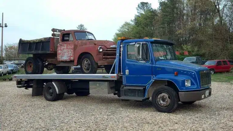 blue rollback tow truck towing an old junk dump truck
